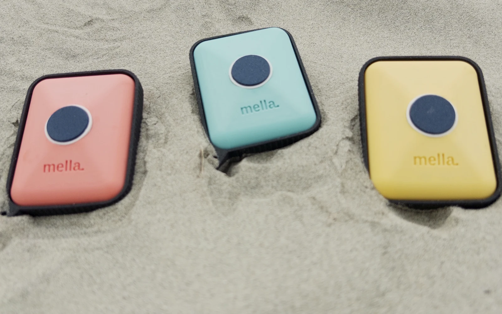 Three Mella prototypes - coral, tidepool blue, and citrus yellow - lined up on open sand.