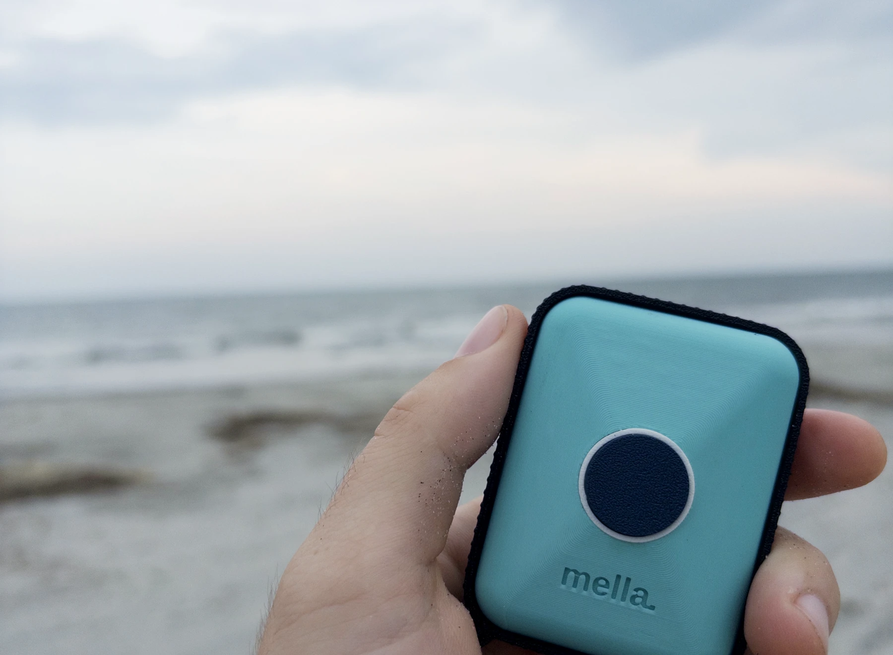 A hand holds the tidepool-blue Mella prototype up against the surf on a Charleston beach.
