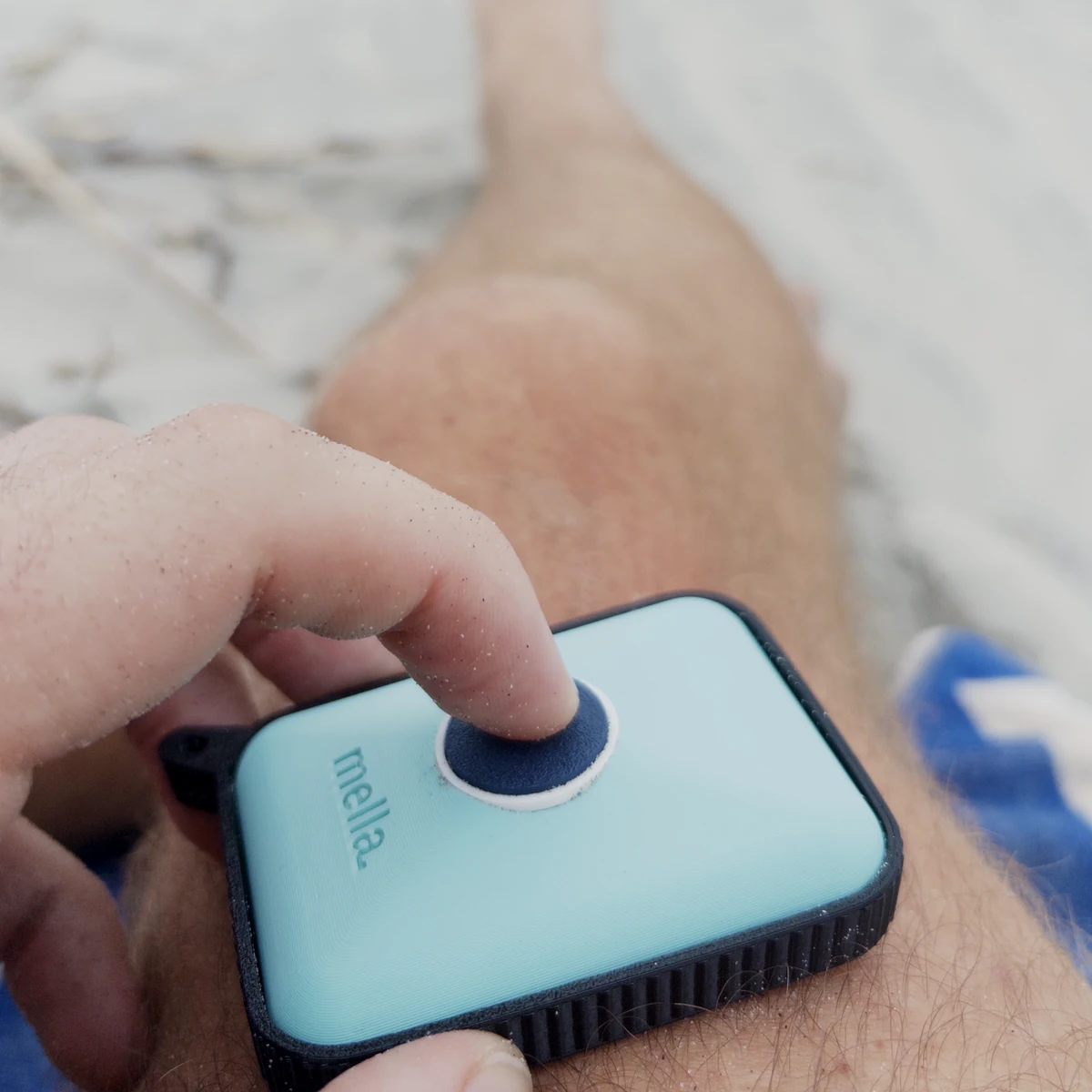 A thumb presses the single navy button on a Mella prototype resting against a leg at the beach.